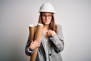 Young beautiful redhead architect woman wearing glasses and safety helmet holding blueprints with surprise face pointing finger to himself