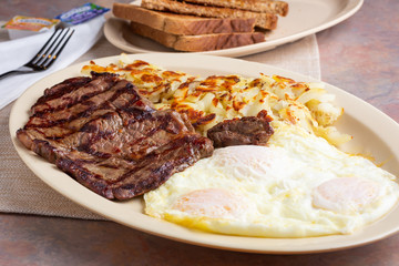 A view of a breakfast plate featuring over easy eggs, a steak cut, hash browns and toast, in a restaurant or kitchen setting.