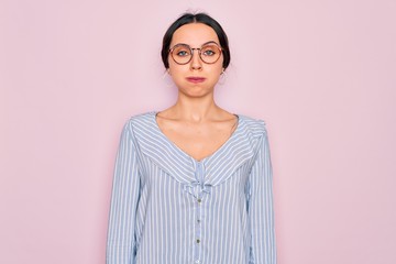 Young beautiful woman wearing casual striped shirt and glasses over pink background puffing cheeks with funny face. Mouth inflated with air, crazy expression.