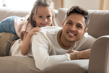 Happy young father lying on comfortable couch with smiling small cute daughter on back. Joyful dad relaxing on sofa with little kid girl, enjoying lazy free weekend time in living room together.