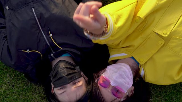 Beautiful Caucasian girls in black and pink medical protective faces masks lie on green grass, talk and look at the sky. Woman in a yellow jacket and glasses shows a finger up on a sunny day. Top view