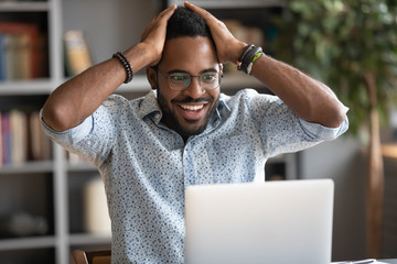 Excited African American man in glasses triumph winning online lottery on laptop, overjoyed...
