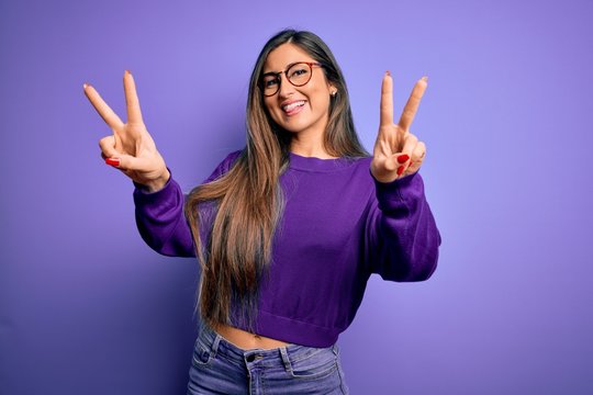 Young beautiful smart woman wearing glasses over purple isolated background smiling with tongue out showing fingers of both hands doing victory sign. Number two.