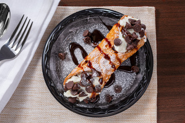 A top down view of a cannoli on a saucer, in a restaurant or kitchen setting.