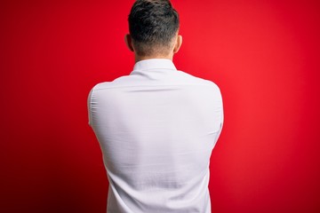 Young business man with blue eyes wearing elegant shirt standing over red isolated background standing backwards looking away with crossed arms