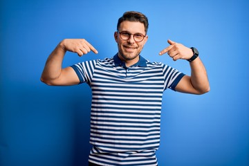 Fototapeta premium Young man with blue eyes wearing glasses and casual striped t-shirt over blue background looking confident with smile on face, pointing oneself with fingers proud and happy.