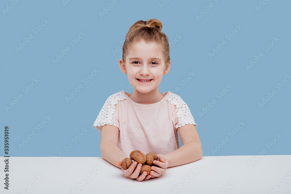 Wall mural portret of little girl with walnuts in hand isolated on blue background, sitting at a table - Wall murals