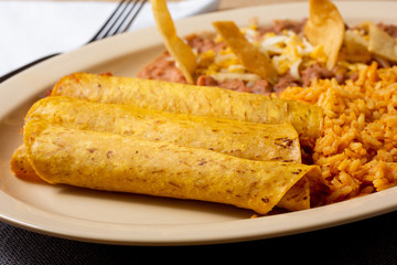 A closeup view of a taquito entree, with sides of rice and beans, in a restaurant or kitchen setting.
