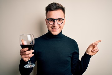 Young handsome caucasian man drinking an alcoholic glass of red wine over isolated background very happy pointing with hand and finger to the side