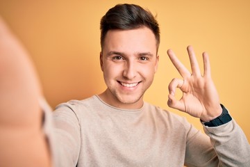 Young handsome caucasian man talking a selfie picture over yellow isolated background doing ok sign...