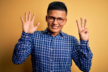 Young handsome latin man wearing casual shirt and glasses over yellow background showing and pointing up with fingers number eight while smiling confident and happy.