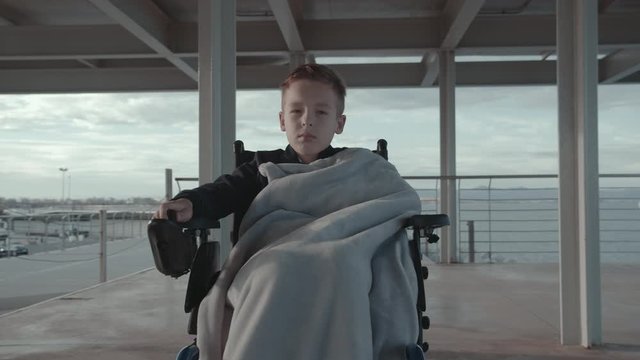 Front view of serious and sad handicapped boy moving in electric wheelchair. He spending time outside on cool day and covered with warm blanket