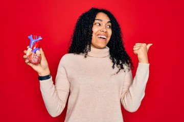 Young african american curly woman holding plastic heart standing over red background pointing and showing with thumb up to the side with happy face smiling