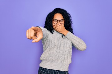 Young african american woman wearing casual sweater and glasses over purple background laughing at you, pointing finger to the camera with hand over mouth, shame expression