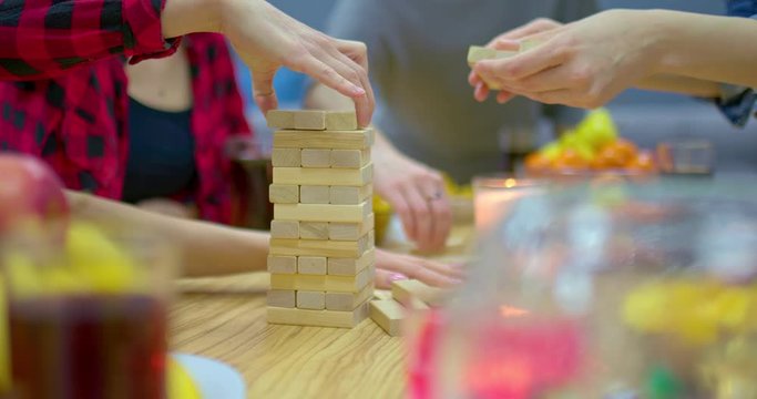 Friends play jenga, stack the bars in the tower. Hands take the bars and lay out the tower. Company plays board games. Close-up.