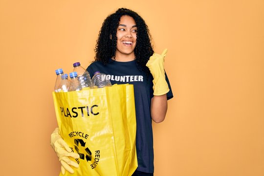 Young african american woman doing volunteering recycling holding bag with plastic bottles pointing and showing with thumb up to the side with happy face smiling