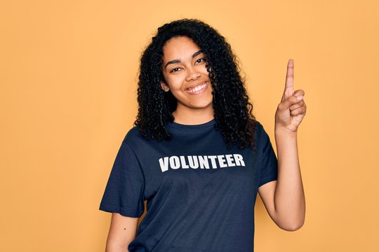Young african american curly woman doing volunteering wearing volunteer t-shirt surprised with an idea or question pointing finger with happy face, number one