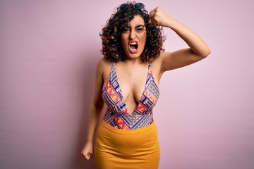 Young beautiful arab woman on vacation wearing swimsuit and sunglasses over pink background angry and mad raising fist frustrated and furious while shouting with anger. Rage and aggressive concept.