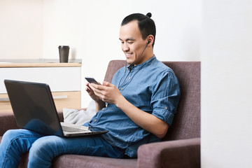 An Asian guy looks at the phone and smiles, works with a laptop in headphones on a sofa at home in the daytime. Remote work outside the office. Hobbies and leisure
