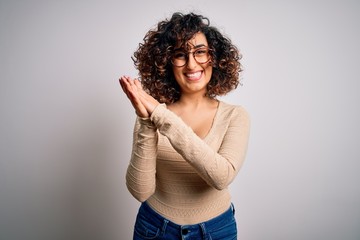 Young beautiful curly arab woman wearing casual t-shirt and glasses over white background clapping...