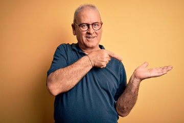 Middle age handsome hoary man wearing casual polo and glasses over yellow background amazed and smiling to the camera while presenting with hand and pointing with finger.