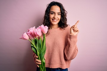 Young beautiful romantic woman with curly hair holding bouquet of pink tulips cheerful with a smile on face pointing with hand and finger up to the side with happy and natural expression