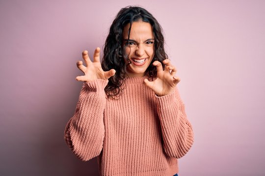 Young beautiful woman with curly hair wearing casual sweater over isolated pink background smiling funny doing claw gesture as cat, aggressive and sexy expression