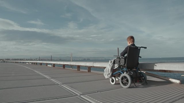 Wide shot of handicapped boy in electric wheelchair riding along the sea and enjoying fresh air