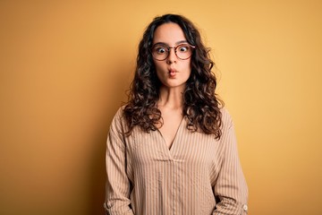 Beautiful woman with curly hair wearing striped shirt and glasses over yellow background making fish face with lips, crazy and comical gesture. Funny expression.