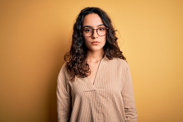 Beautiful woman with curly hair wearing striped shirt and glasses over yellow background Relaxed with serious expression on face. Simple and natural looking at the camera.