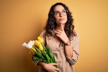 Young beautiful romantic woman with curly hair holding bouquet of yellow tulips Thinking concentrated about doubt with finger on chin and looking up wondering