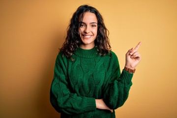 Young beautiful woman with curly hair wearing green casual sweater over yellow background with a big smile on face, pointing with hand and finger to the side looking at the camera.