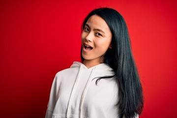 Young beautiful chinese sporty woman wearing sweatshirt over isolated red background winking...