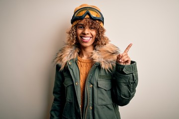 Young african american skier woman with curly hair wearing snow sportswear and ski goggles with a big smile on face, pointing with hand finger to the side looking at the camera.