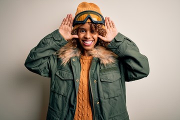 Young african american skier woman with curly hair wearing snow sportswear and ski goggles Smiling cheerful playing peek a boo with hands showing face. Surprised and exited
