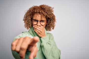 Young beautiful african american woman wearing turtleneck sweater and glasses laughing at you, pointing finger to the camera with hand over mouth, shame expression