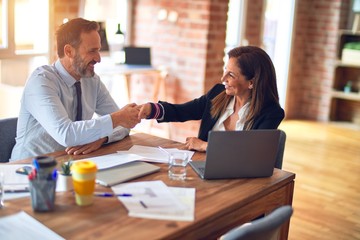 Two middle age business workers smiling happy and confident. Working together with smile on face using laptop doing handsake at the office