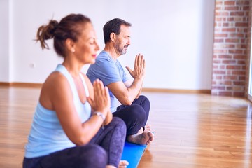 Fototapeta na wymiar Middle age beautiful sporty couple sitting on mat practicing yoga doing prayer pose at gym
