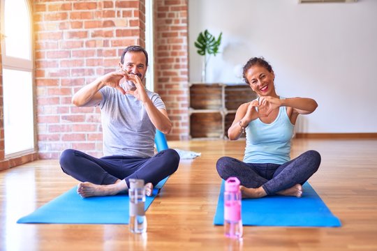 Middle age sporty couple sitting on mat doing stretching yoga exercise at gym smiling in love showing heart symbol and shape with hands. Romantic concept.