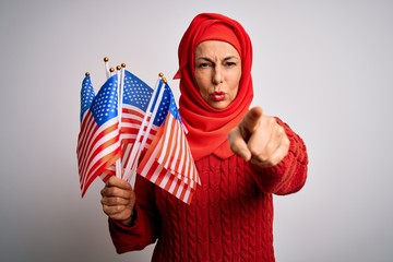 Middle age patriotic woman wearing traditional muslim hijab holding united states flags pointing with finger to the camera and to you, hand sign, positive and confident gesture from the front
