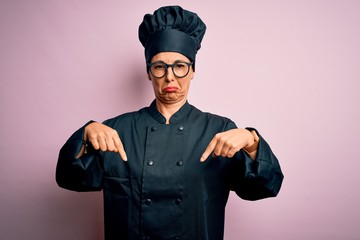 Middle age brunette chef woman wearing cooker uniform and hat over isolated pink background Pointing down looking sad and upset, indicating direction with fingers, unhappy and depressed.