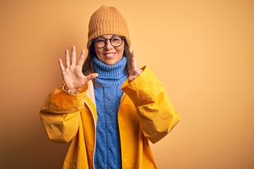 Middle age woman wearing yellow raincoat and winter hat over isolated background showing and pointing up with fingers number six while smiling confident and happy.