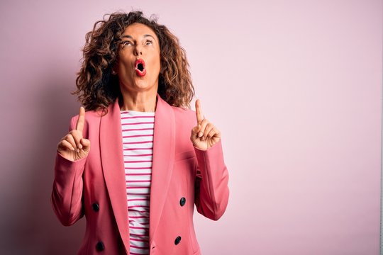 Middle age beautiful businesswoman wearing elegant jacket over isolated pink background amazed and surprised looking up and pointing with fingers and raised arms.