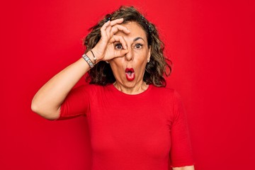 Middle age senior brunette woman wearing casual t-shirt standing over red background doing ok gesture shocked with surprised face, eye looking through fingers. Unbelieving expression.