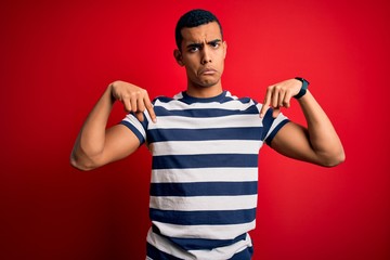 Handsome african american man wearing casual striped t-shirt standing over red background Pointing down looking sad and upset, indicating direction with fingers, unhappy and depressed.