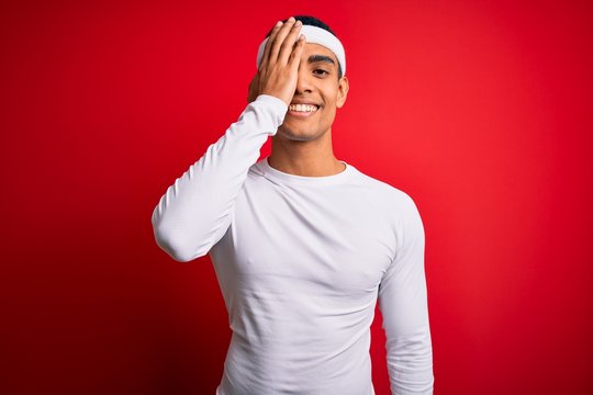 Young handsome african american sportsman wearing sportswear over red background covering one eye with hand, confident smile on face and surprise emotion.