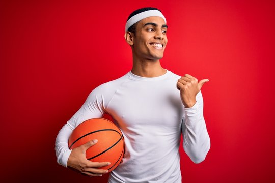 Young handsome african american sportsman holding basketball ball over red background pointing and showing with thumb up to the side with happy face smiling