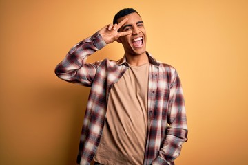 Young handsome african american man wearing casual shirt standing over yellow background Doing peace symbol with fingers over face, smiling cheerful showing victory