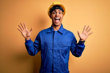 Young handsome african american worker man wearing blue uniform and security helmet celebrating crazy and amazed for success with arms raised and open eyes screaming excited. Winner concept