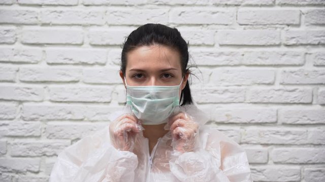 Beautiful European woman with black hair in a white protective suit and gloves on brick wall background puts respirator, glasses and two medical masks on her face, zips jacket, put on a hood close up